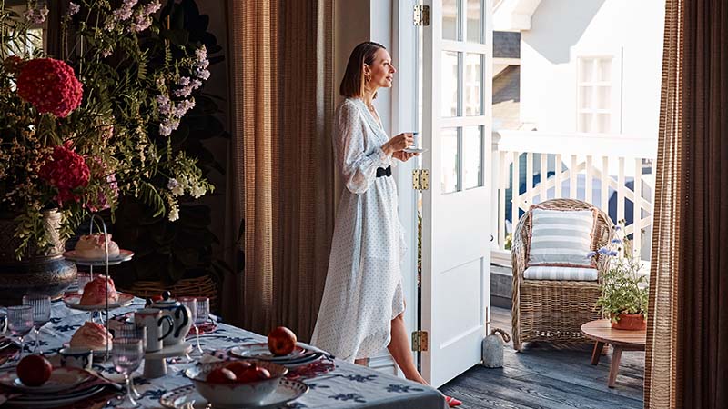 Woman in a white dress holds a cup, standing near an open patio door. A set table with flowers and red fruit is in the foreground, evoking a serene morning.