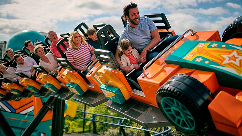 A joyful group of people, including a man and child, ride a colorful orange roller coaster under a blue sky, conveying excitement and fun.