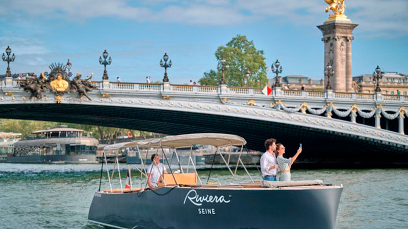 A couple takes a selfie on a small boat labeled "Riviera Seine" beneath a decorative bridge in Paris.