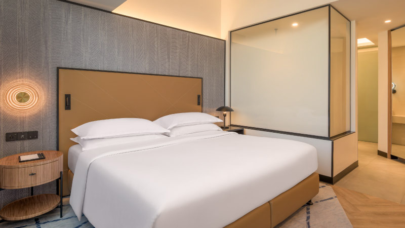 Modern hotel room with a large, neatly made bed with white linens. A wooden nightstand beside it holds a lamp. Glass panel partially divides the room.