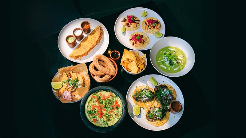 Assorted Mexican dishes including tacos, quesadilla, guacamole, tortilla chips, churros, and soup on a dark table