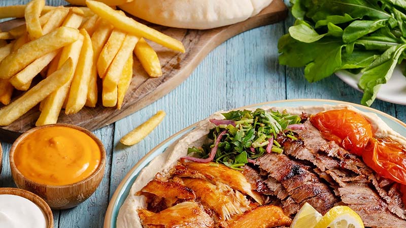 Mixed shawarma plate with sliced beef and chicken, fries, sauce, pita bread, and fresh greens