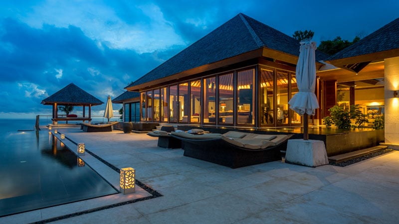 Luxurious beachfront villa at dusk, featuring large, illuminated windows, lounge chairs by an infinity pool, and a serene, inviting atmosphere.