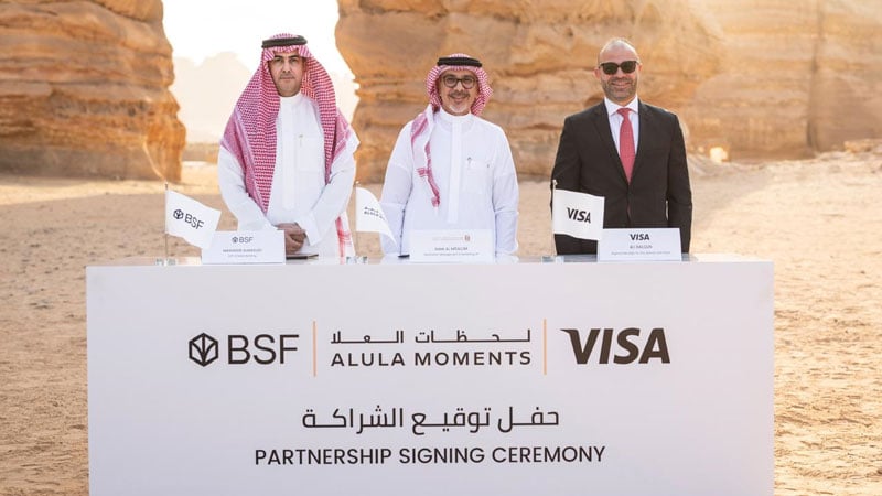 Three men stand behind a partnership signing ceremony table in a desert setting. The table displays logos for BSF, AlUla Moments, and Visa.