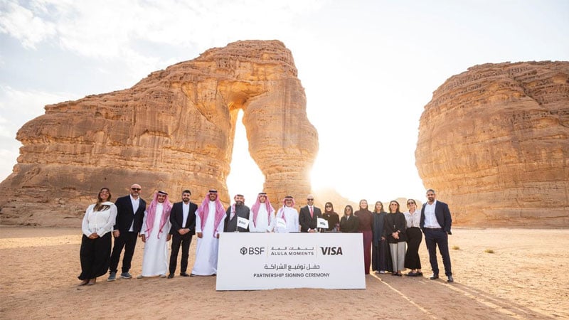 A group of people stands in front of a large rock formation at sunset. They are dressed formally, gathered around a sign for a partnership signing ceremony.