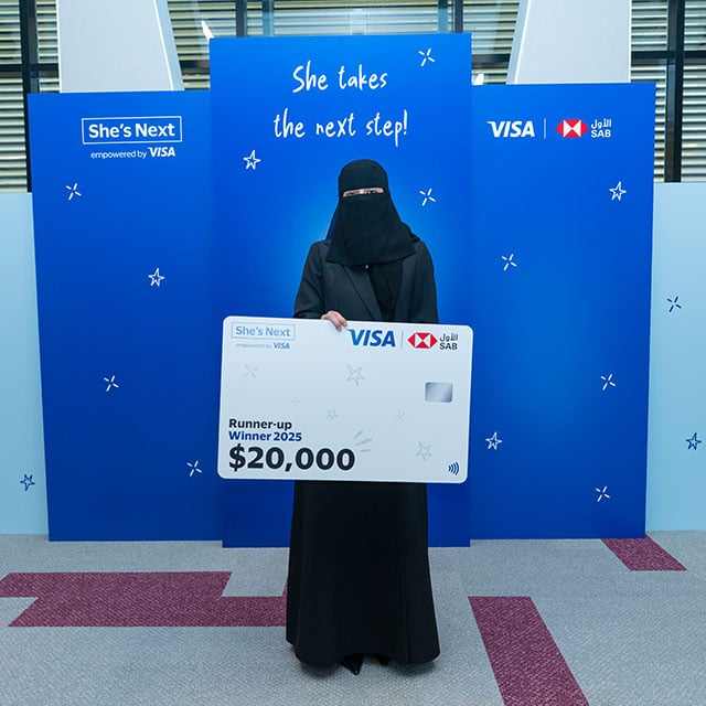 A woman dressed in black holds a large check for $20,000, standing in front of a blue backdrop with "She takes the next step!" written on it.