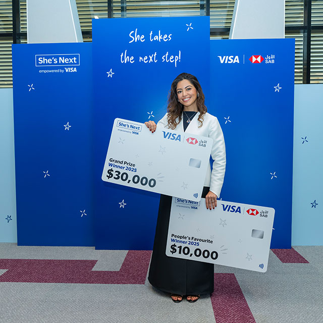 A woman joyfully holds two large mock credit cards displaying prize amounts of $30,000 and $10,000. She stands in front of a blue backdrop with "She takes the next step!" written on it, alongside Visa and SAB logos. The scene conveys excitement and achievement.